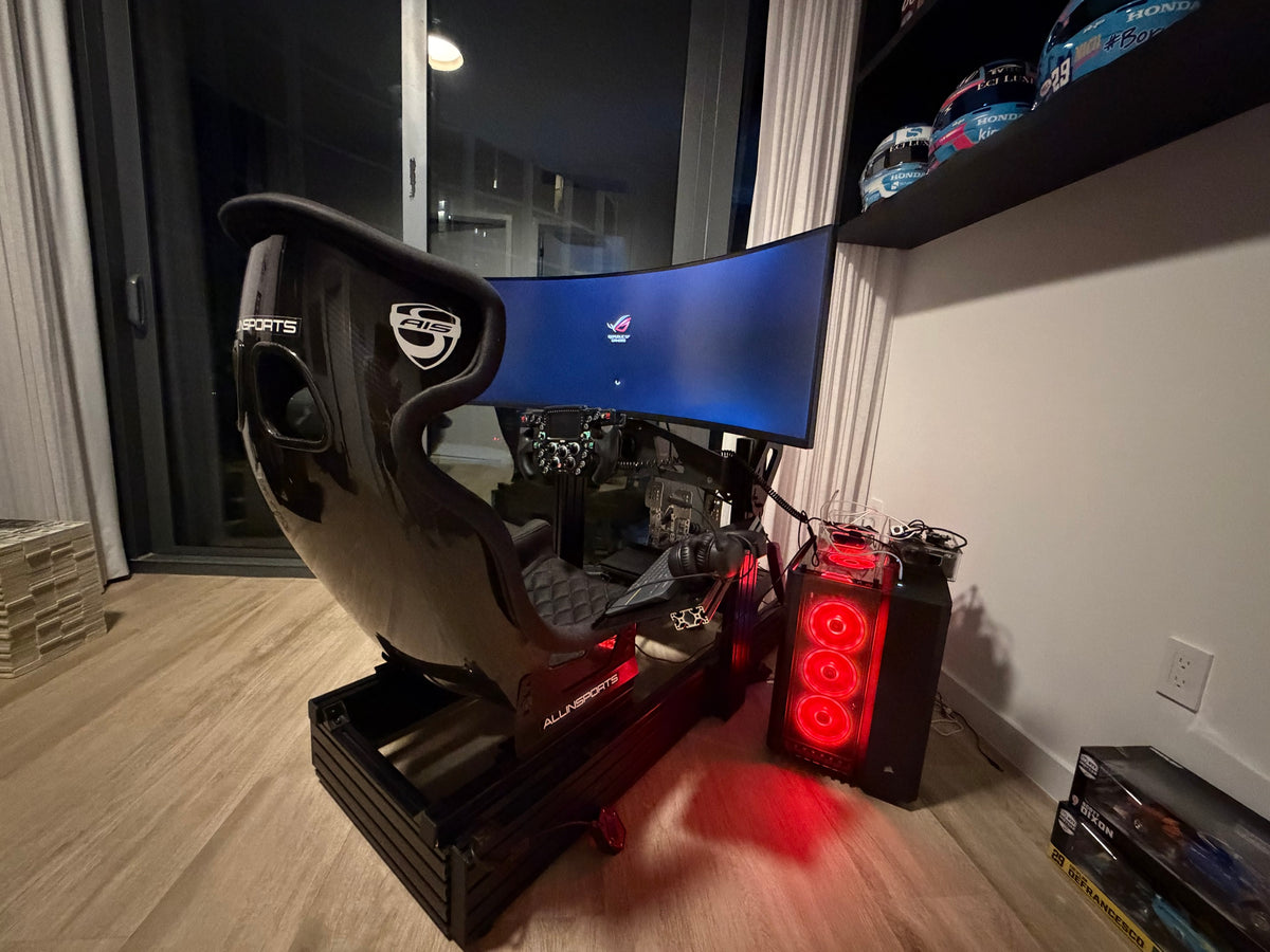 Completed sim racing build with a carbon shell seat, formula wheel and curved ultrawide screen, race helmets on the shelf above